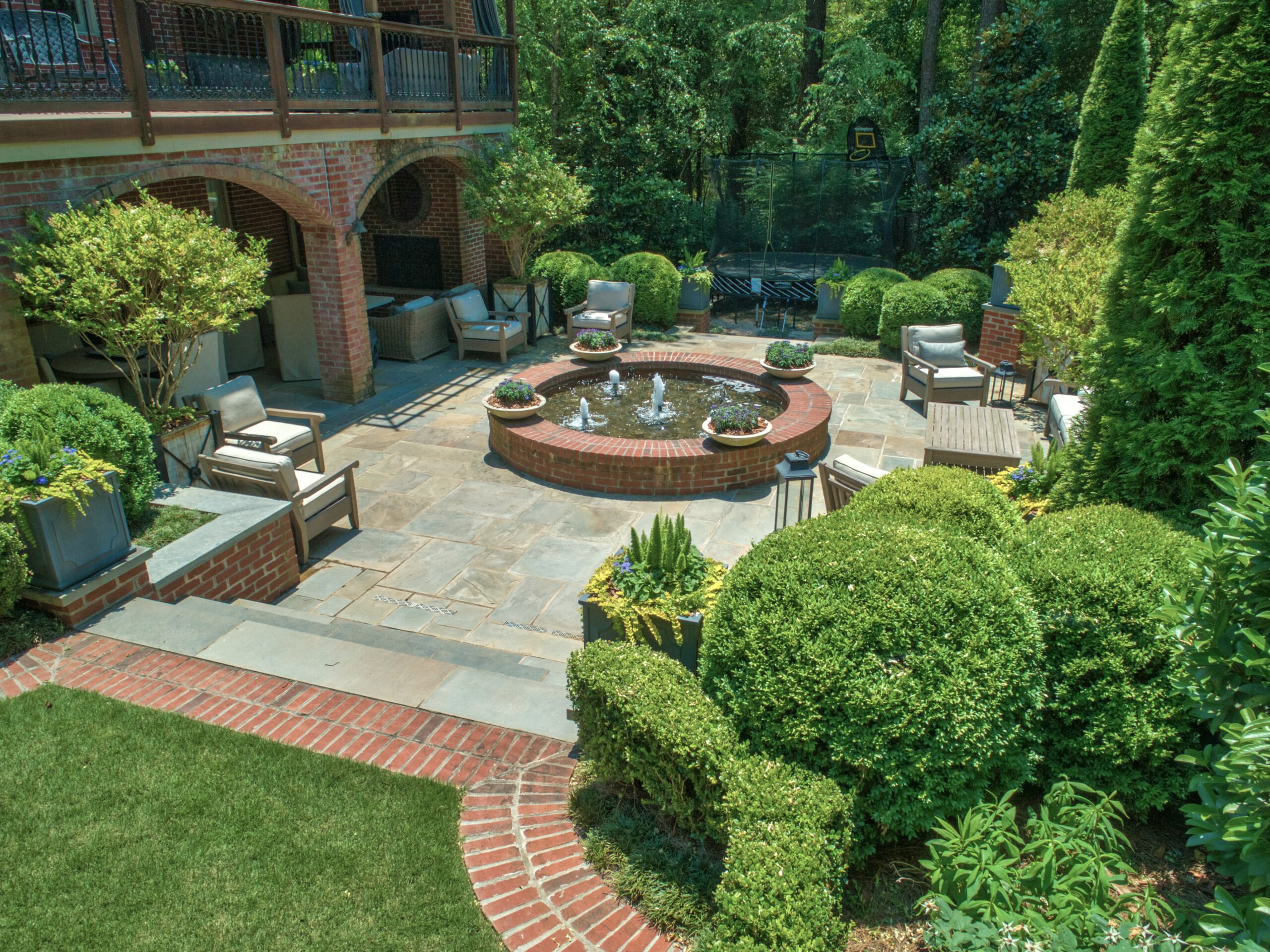 A view of a backyard paved with stone slabs around a brick fountain with a trampoline hidden in the greenery