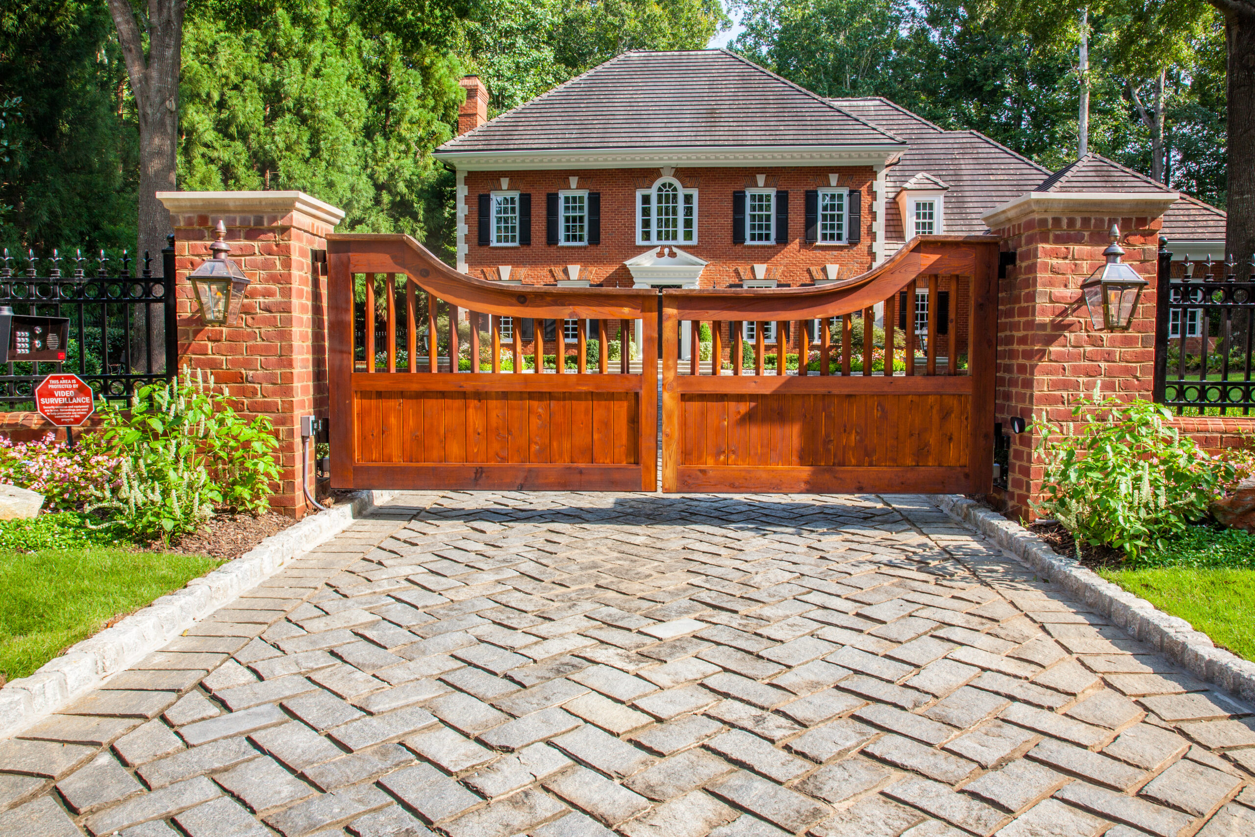 A cedar gate connected to a brick wall in front of a brick house over a herringbone stone brick driveway