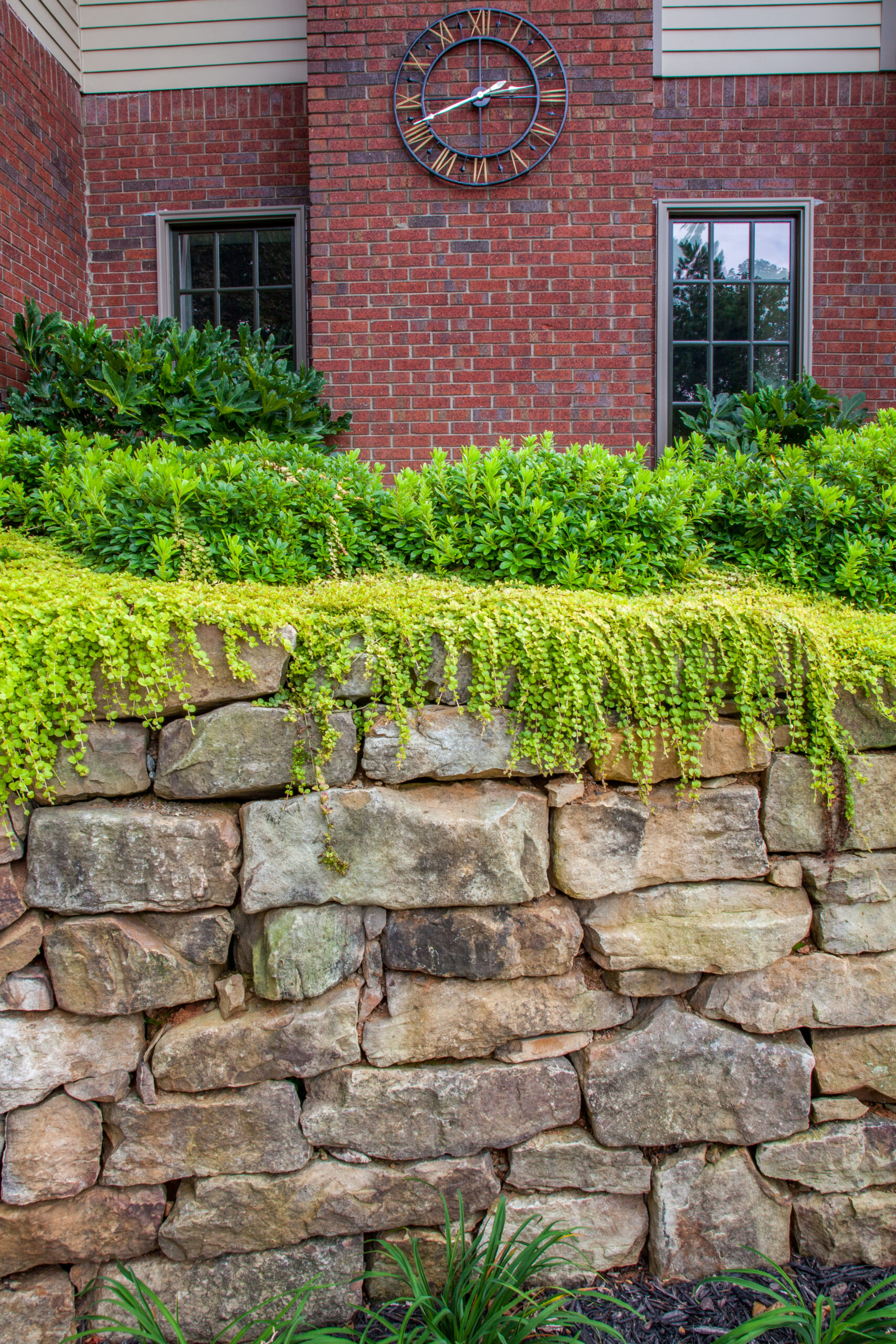 A retaining wall in front of a brick house with green landscaping and a clock on the side of the house