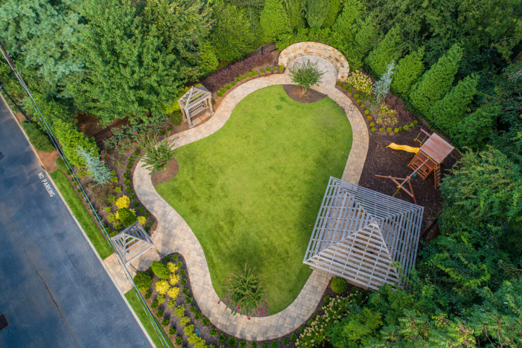 An overhead shot of a small park with a grassy green lawn surrounded by a stone path with natural wood arbors and a play structure