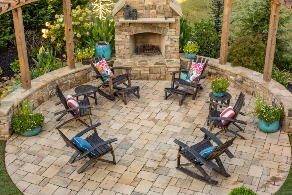 a stone patio with an outdoor fireplace and dark stained Adirondack chairs surrounded by greenery