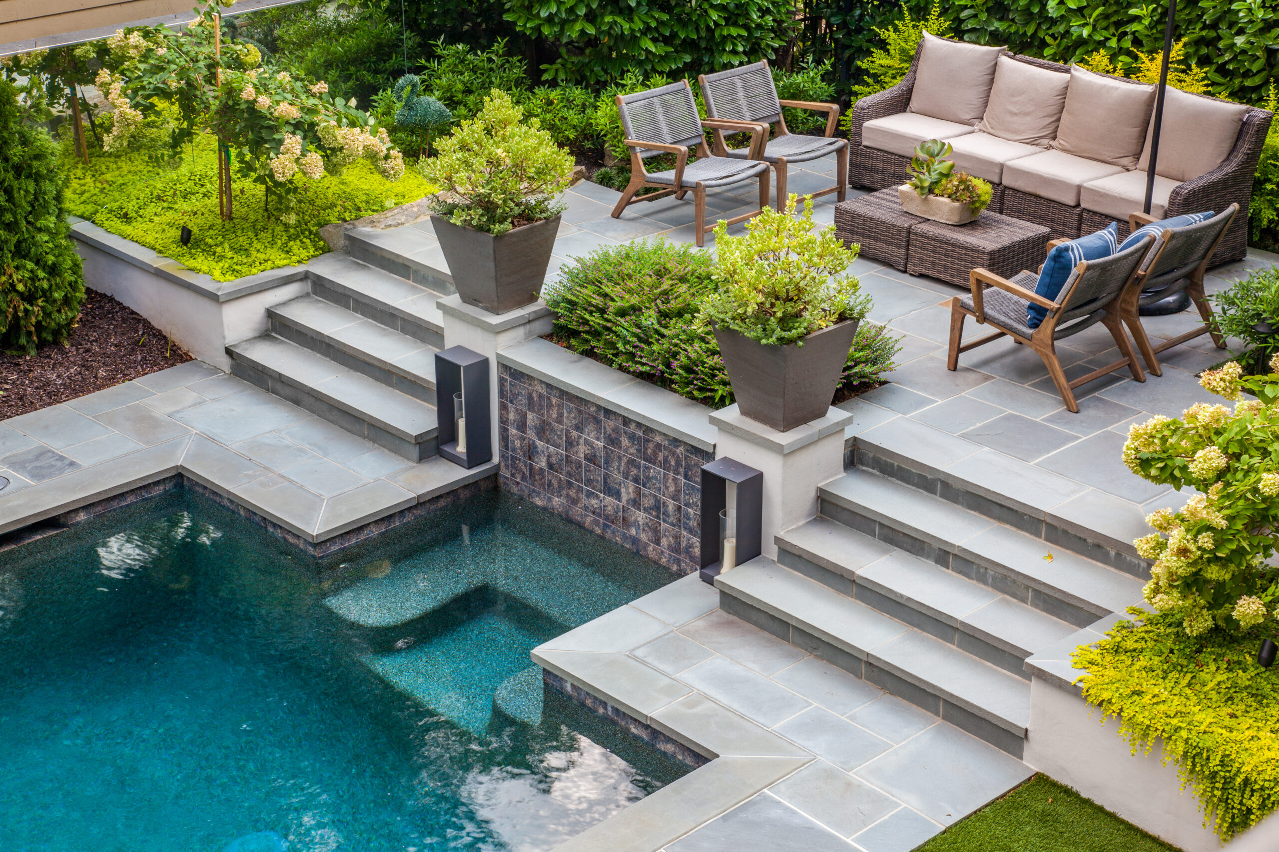 an overhead shot of an outdoor seating area on a raised terrace paved with stone slabs a few steps above a custom pool