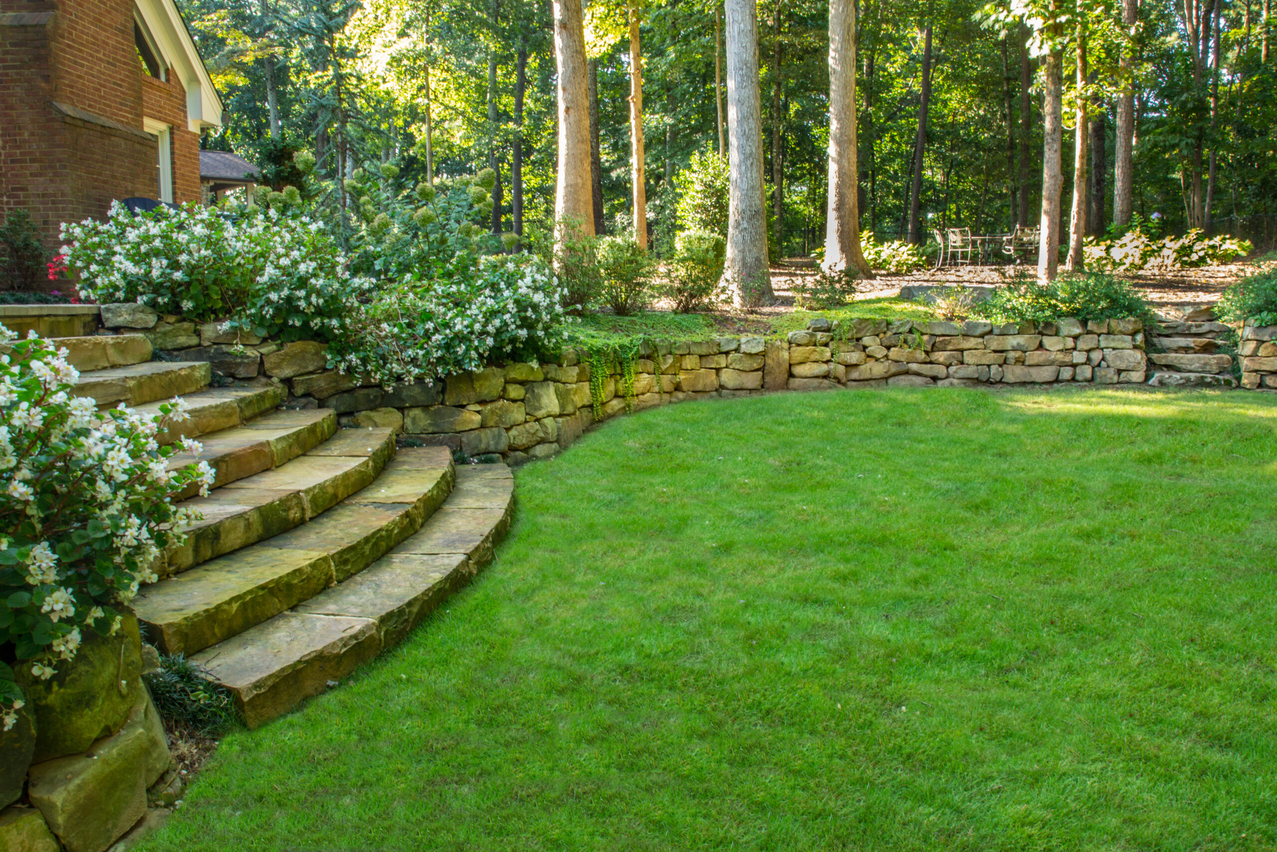 a gorgeous backyard with a lush grass terrace, stone retaining wall, and greenery