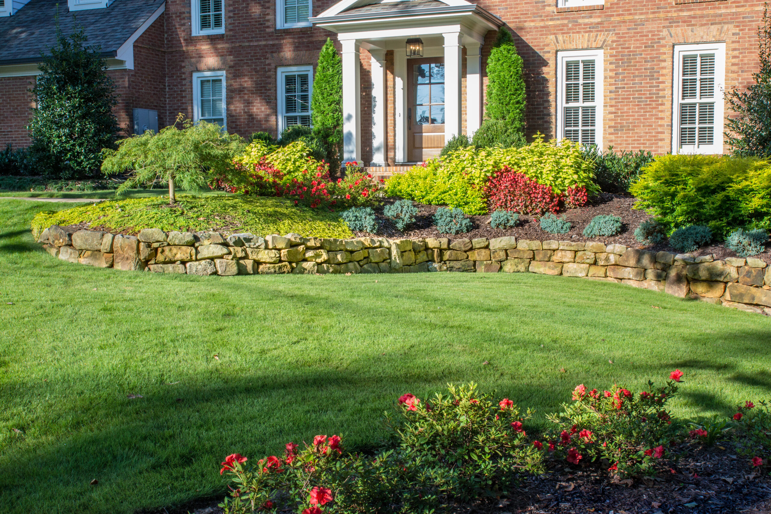 a gorgeous retaining wall at the front of a brick house landscaped with green and red plants