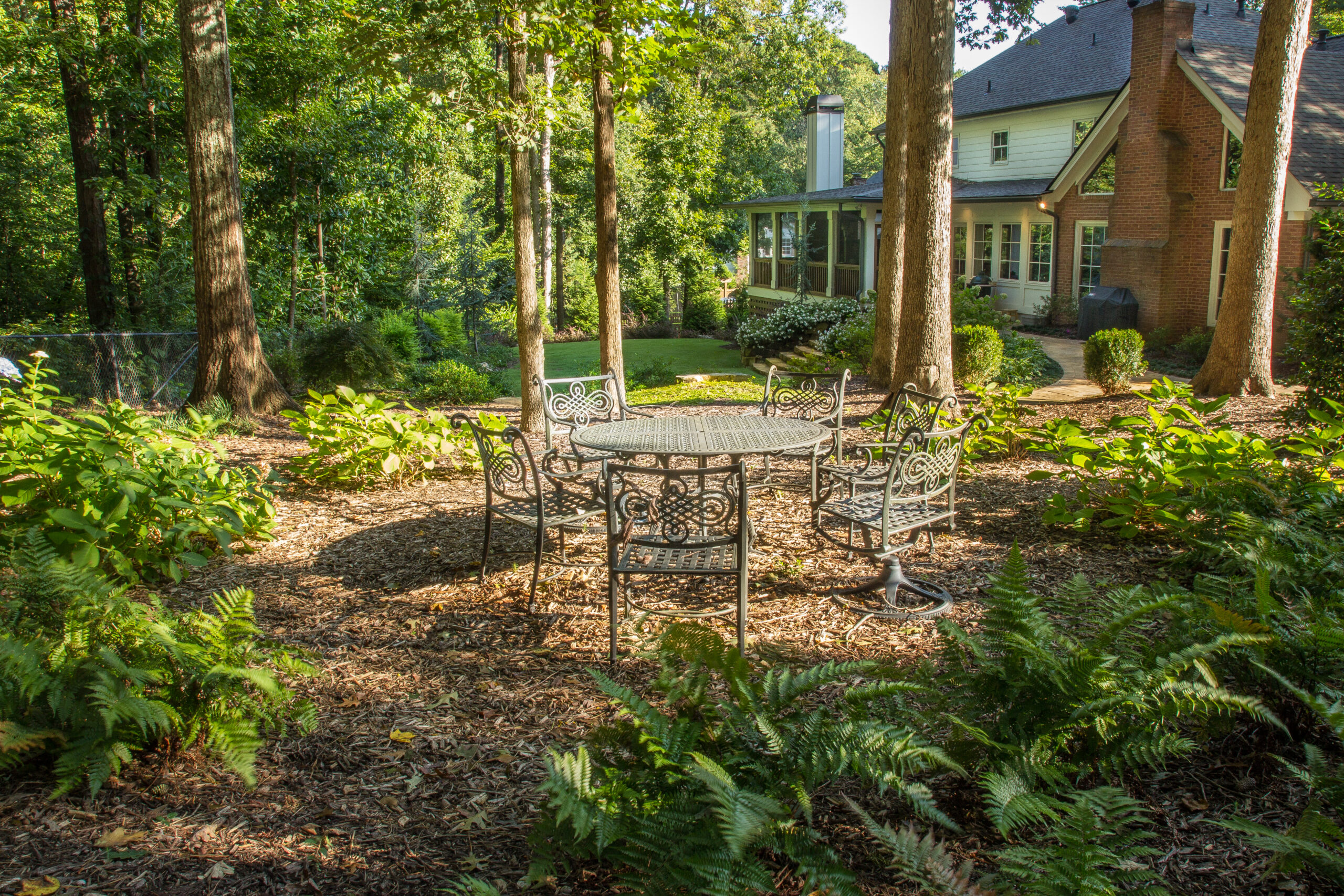 A wrought iron round table with six chairs surrounded by trees in the backyard of a brick house