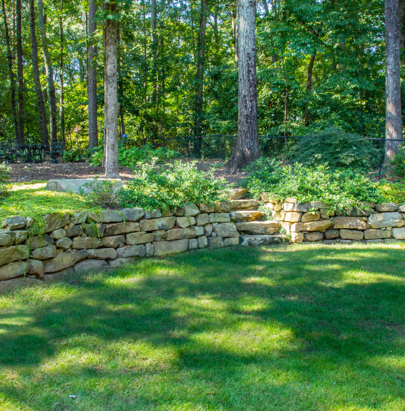 a grassy backyard with stone brick steps leading up to an iron table surrounded by trees