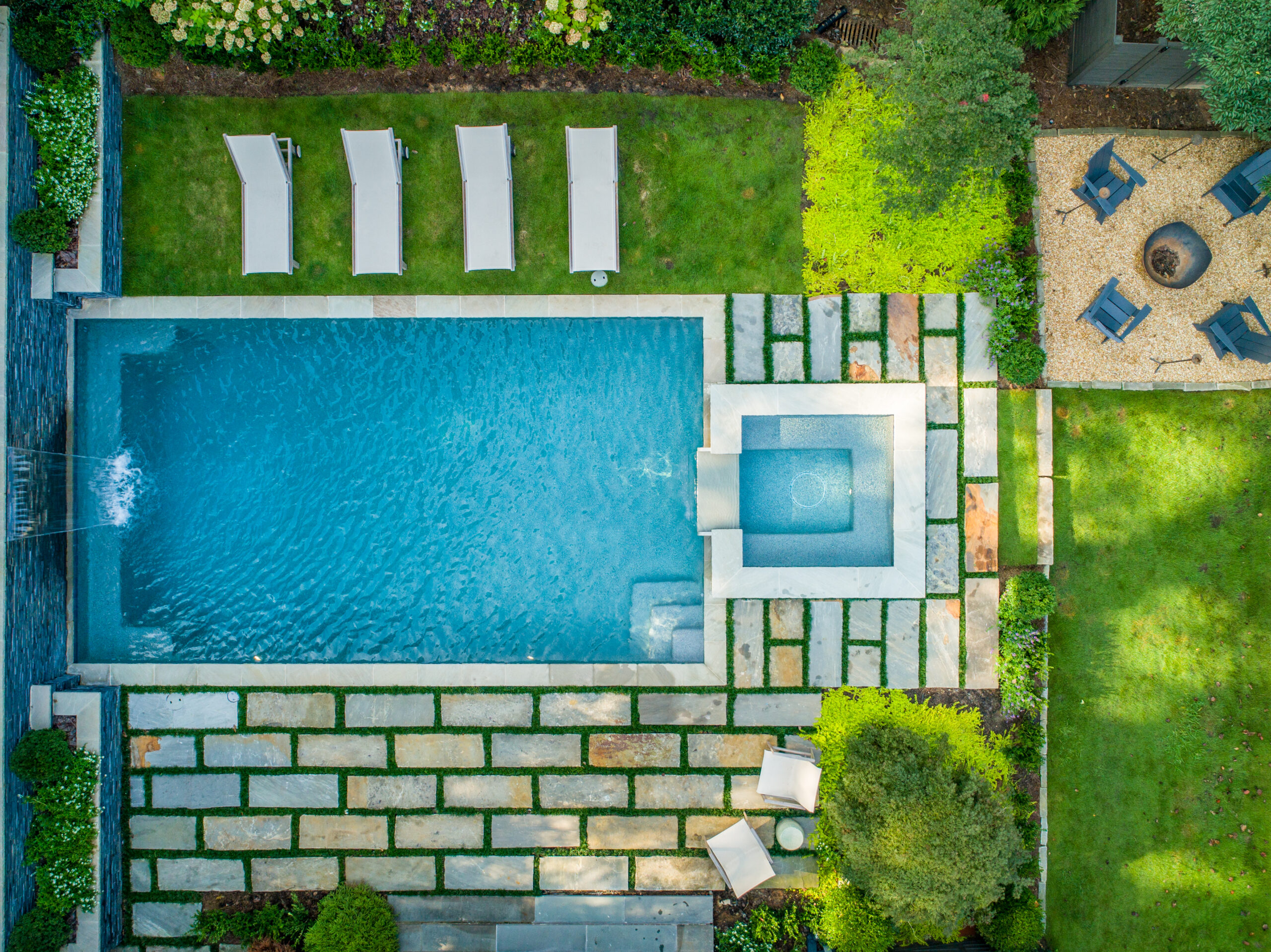 an overhead shot of a pool and spa with greenery surrounding the backyard and four chairs surrounding a fire pit in the corner