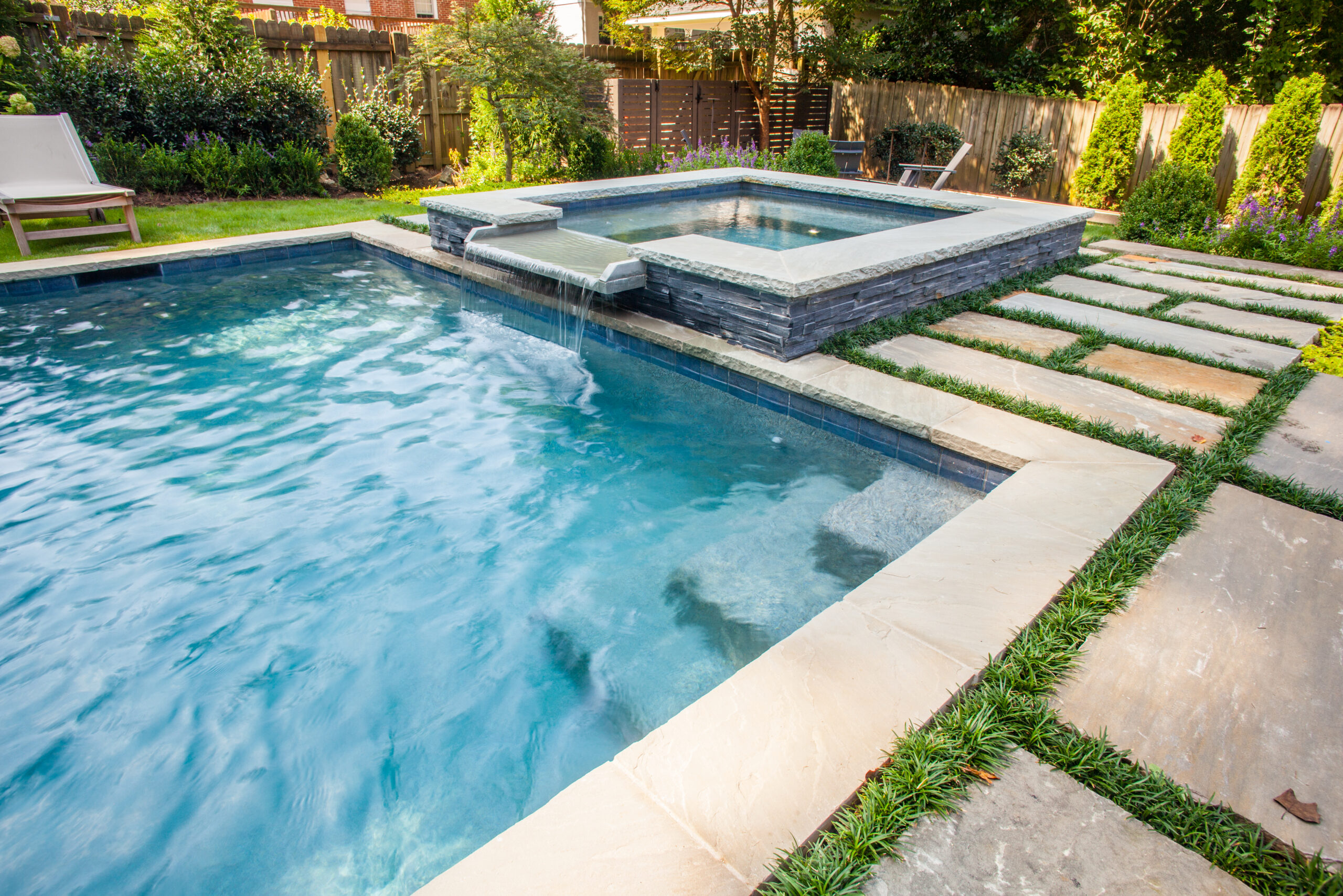 an in-ground spa with a spillover fountain going into a pool surrounded by rectangular pavers with grass between them