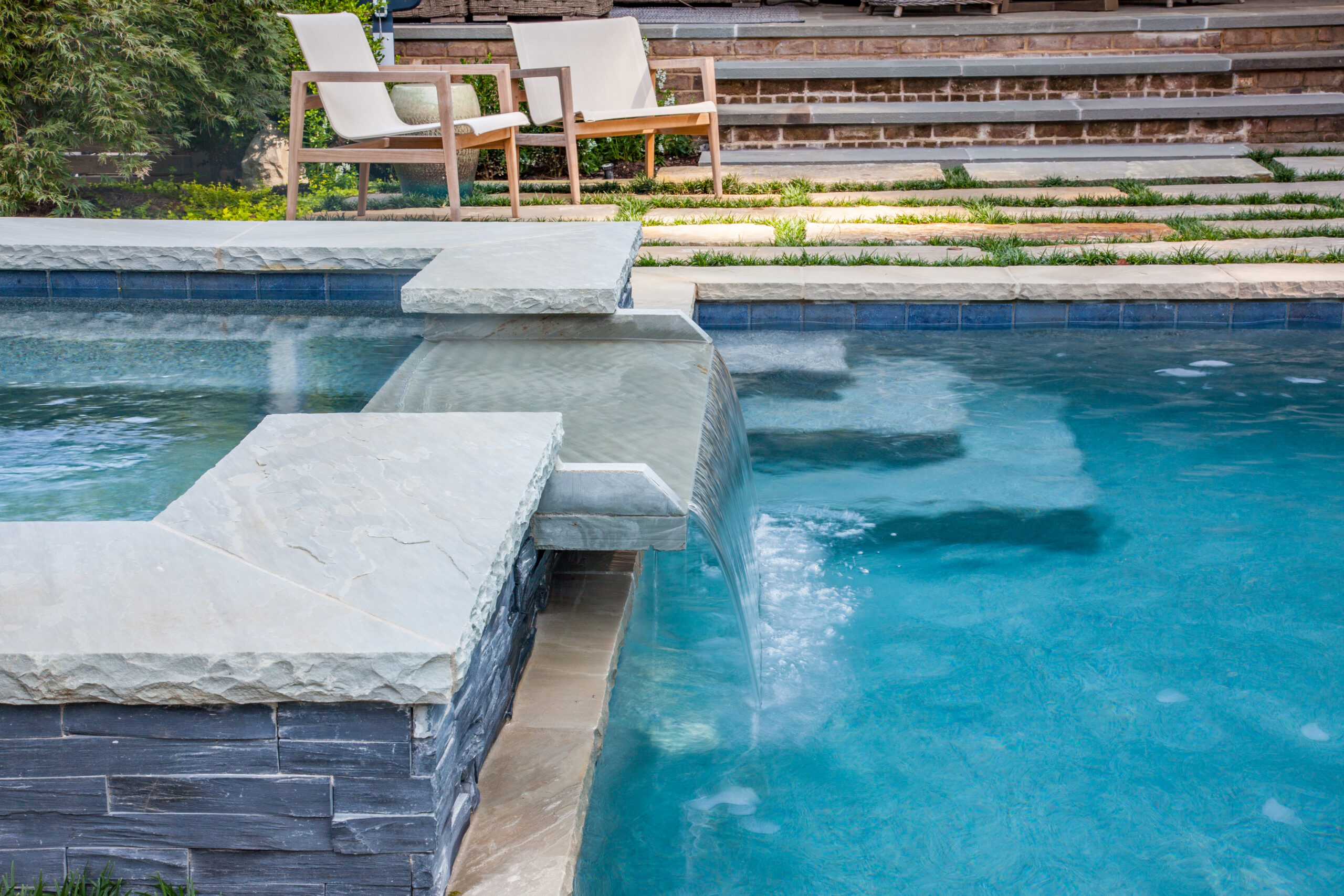 a spillover fountain from an in-ground spa pouring into a swimming pool with lounge chairs in the background