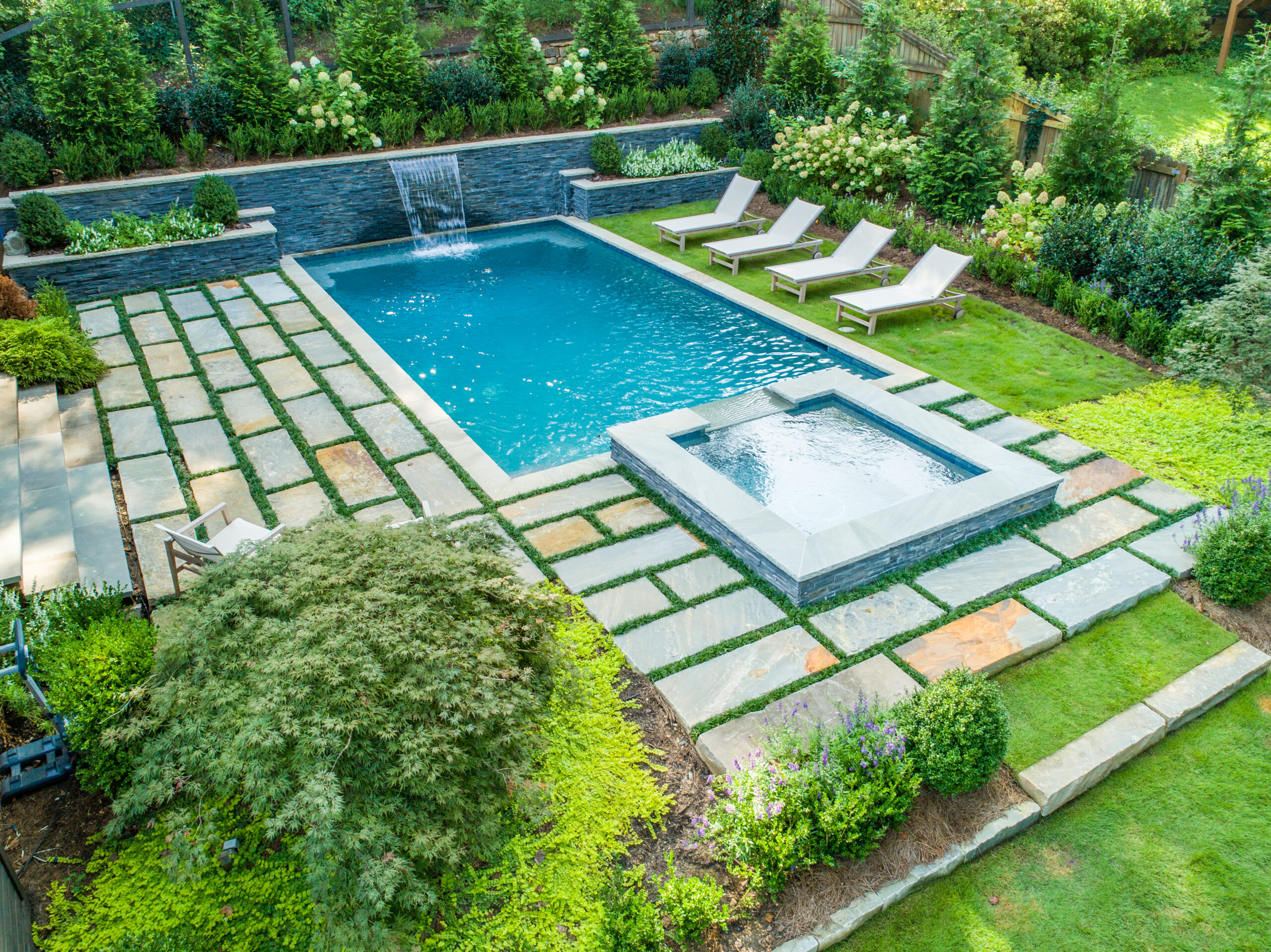 a drone shot of an in-ground pool and spa with lounge chairs on grass and rectangular pavers with grass between them surrounded by greenery