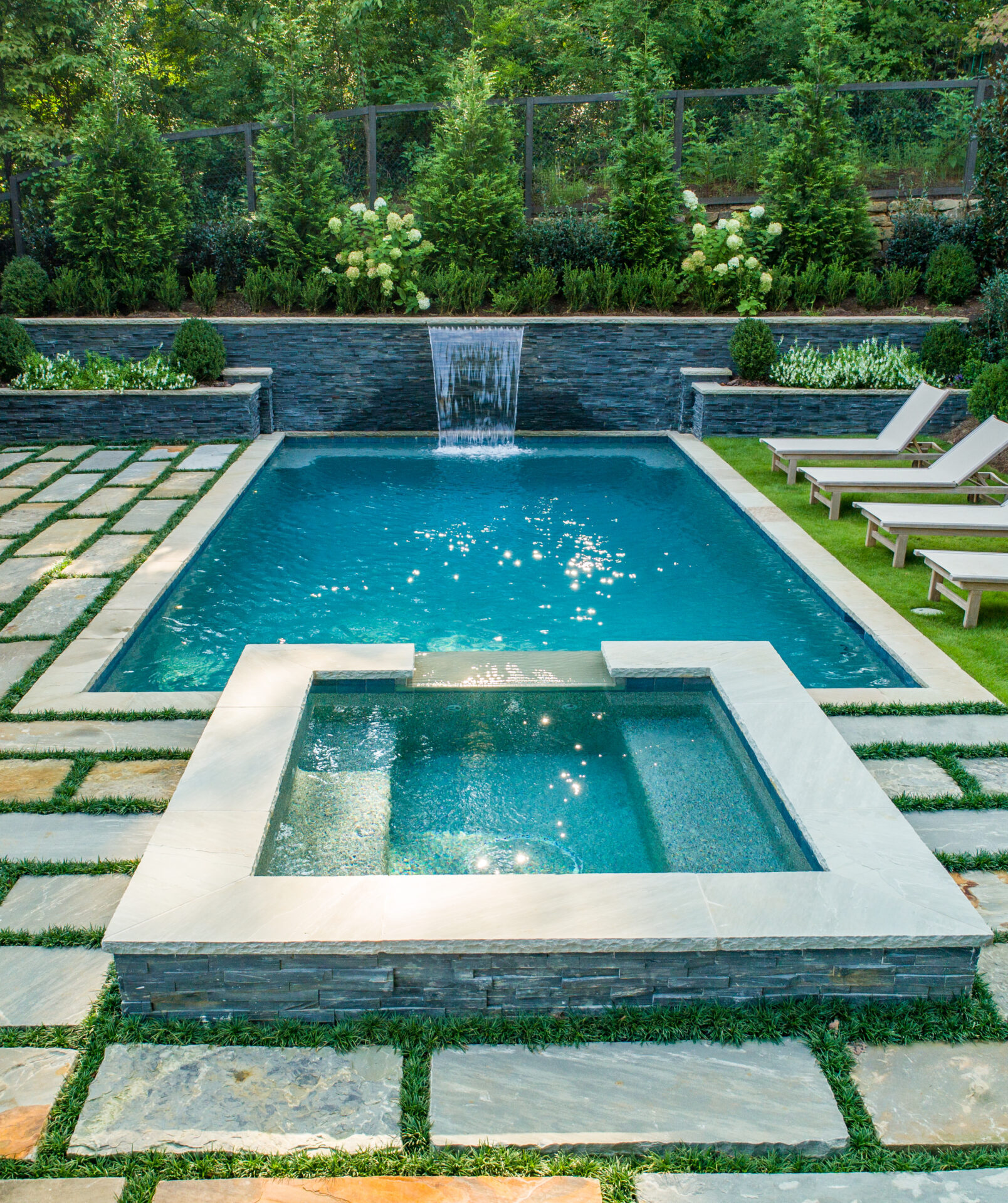 a beautiful pool with a fountain and attached spa surrounded by stone pavers with ornamental grass and modern lawn chairs