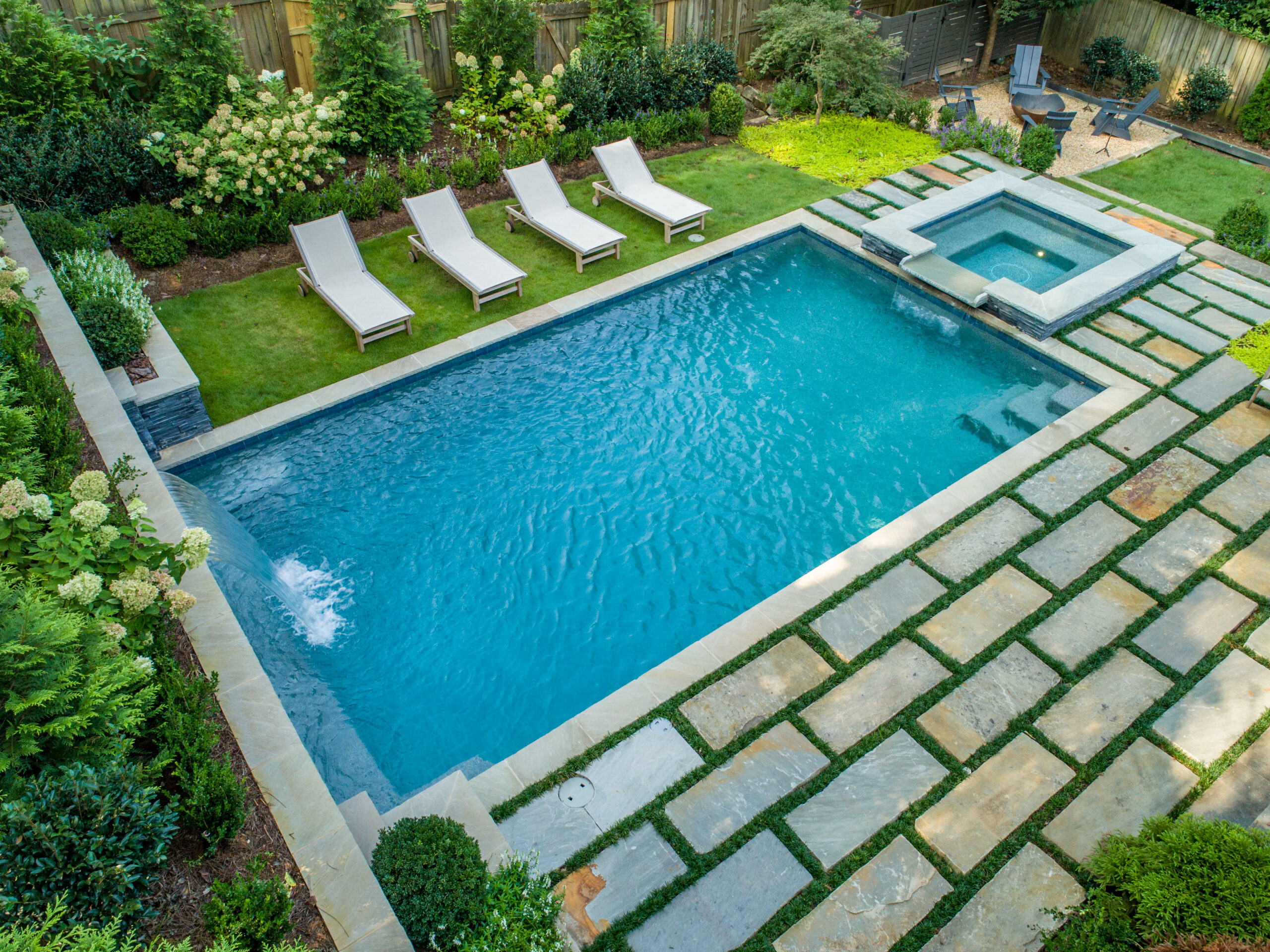 an overhead shot of a beautiful backyard complete with a pool and attached spa, firepit, lawn chairs and surrounded by greenery