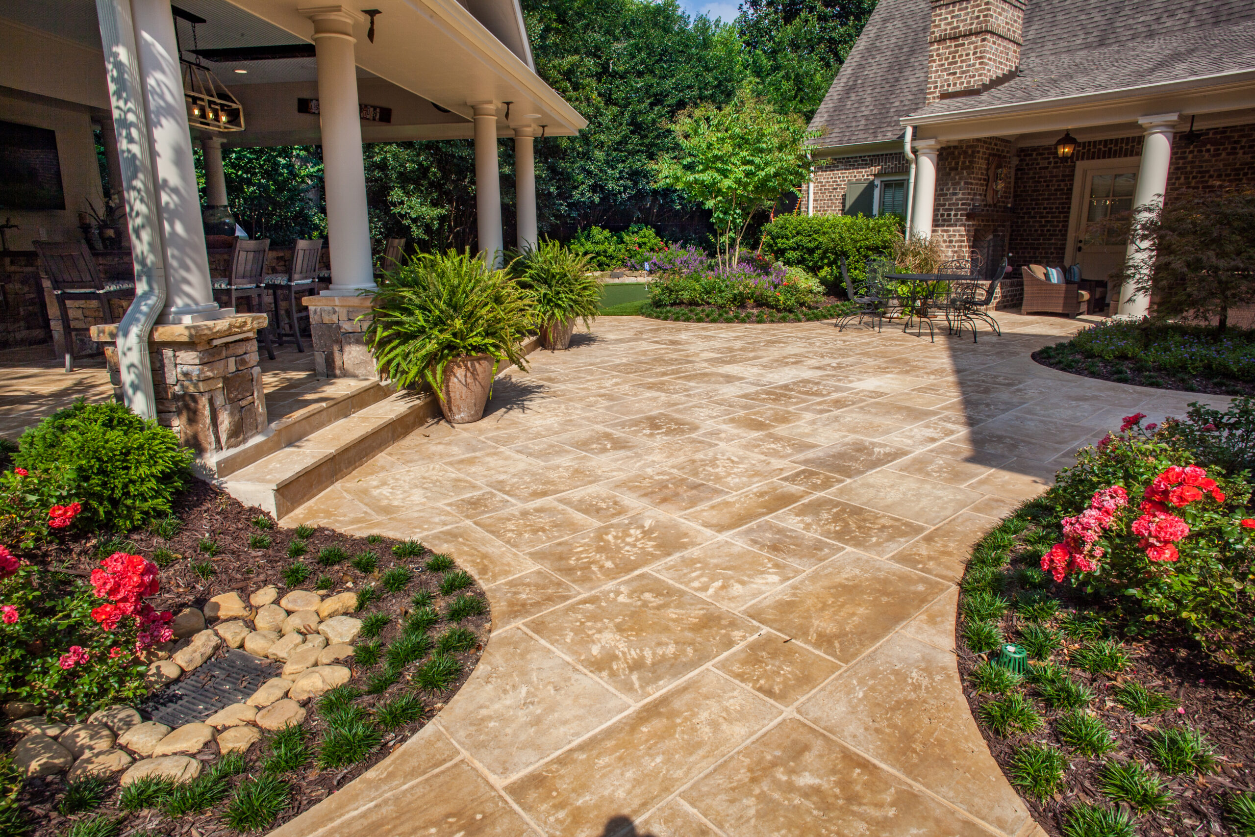 A stone-paved patio between a brick house and a luxurious outdoor kitchen surrounded by greenery