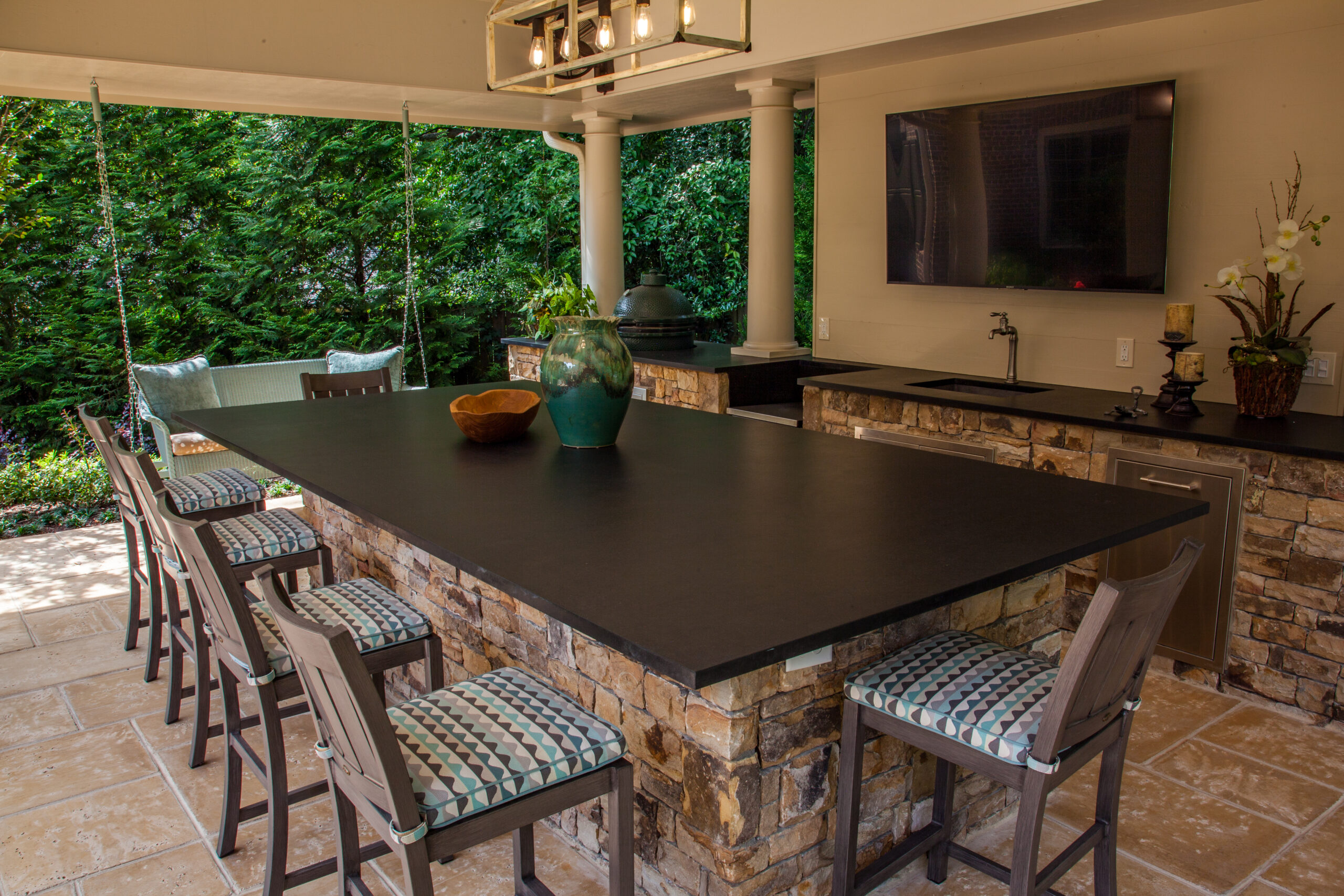 a covered outdoor kitchen with a large tv, seating around a kitchen island, and a swinging bench in the background