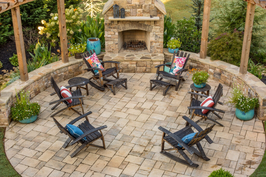 Professionally designed backyard patio with a circular paver seating area, natural stone outdoor fireplace, Adirondack chairs, and lush landscaping, creating a warm and inviting outdoor living space.